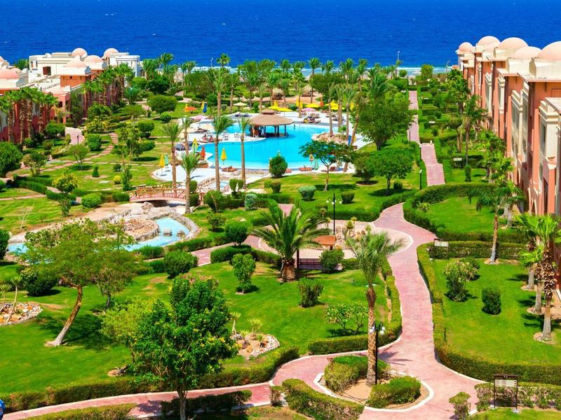 Große Gartenanlage mit Palmen, Pool und Wegen, umgeben von roten Gebäuden am Meer.