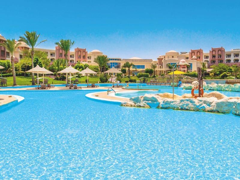 Großer blauer Hotelpool mit Liegestühlen, Sonnenschirmen, Palmen und bunten Gebäuden im Hintergrund.