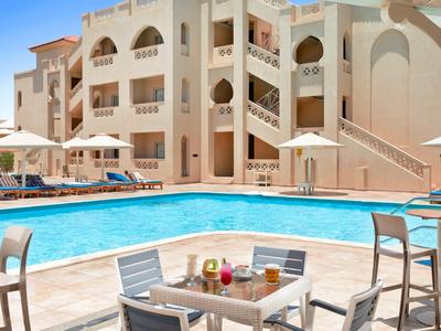 Nowoczesny basen hotelowy z leżakami, stołami i parasolami przed wielopiętrowym budynkiem.