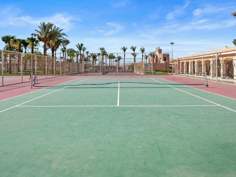 Leerer Tennisplatz mit grünem Spielfeld, umgeben von roten Linien und Palmen unter blauem Himmel.