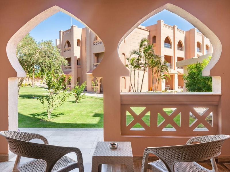 Blick durch bogenförmige Fenster auf Garten, Pflanzen und Gebäude, mit zwei Stühlen und Tisch davor.