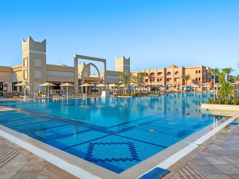 Großer blauer Hotelpool mit Palmen und sandfarbenen Gebäuden unter klarem Himmel.