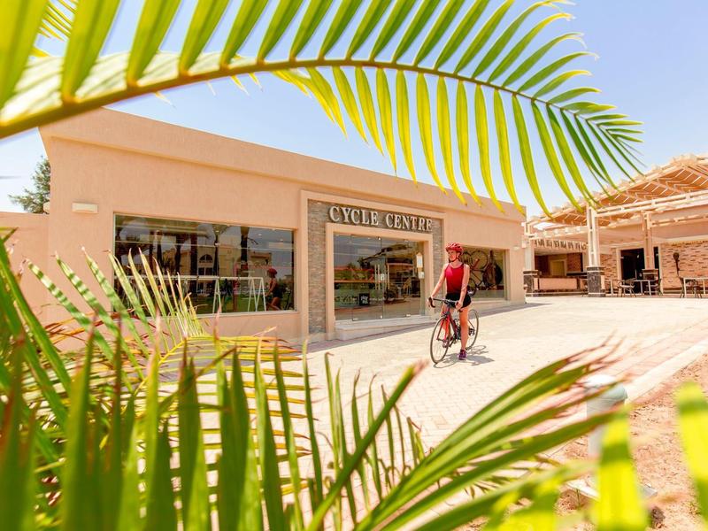 Eingang des Einkaufszentrums "Cala D'Or" mit Radfahrer, Palmenblättern und klarem Himmel.
