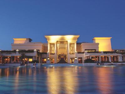 Großes, beleuchtetes Gebäude mit Säulen, davor ruhiger Wasserpool, blauer Abendhimmel.