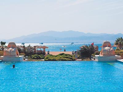 Großer blauer Pool mit Palmen und Meer im Hintergrund, bedeckter Himmel, Insel am Horizont.
