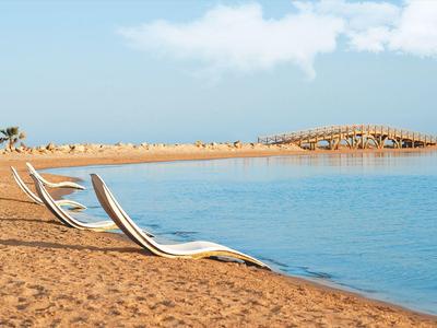 Twee ligstoelen op een zandstrand naast kalm blauw water onder een halfbewolkte lucht.