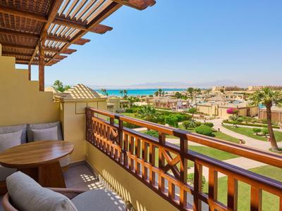Balkon mit Holzmöbeln, Blick auf Garten, Palmen und das blaue Meer am klaren Himmel.