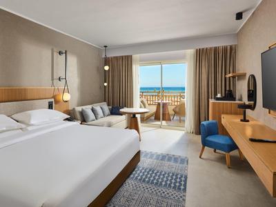 Modernes Hotelzimmer mit großem Bett, Sofa, TV, Schreibtisch und Balkon mit Meerblick.