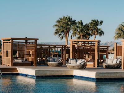 Moderner Hotelpool mit Holzpavillons und Palmen im Hintergrund bei klarem Himmel.