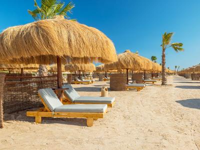 Reihen von Liegestühlen mit Strohdächern am sonnigen Sandstrand eines Urlaubsresorts.