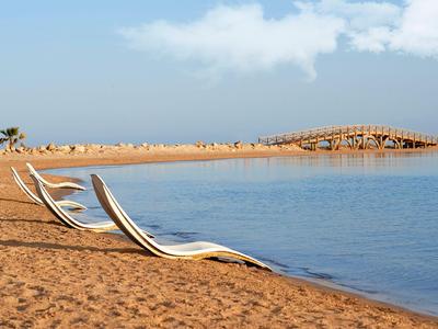 Leere Liegestühle am ruhigen Strand mit Blick auf das blaue Wasser und einen Steg.