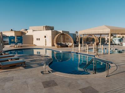 Moderner Hotelpool mit Liegestühlen und überdachten Sitzbereichen bei klarem blauem Himmel.