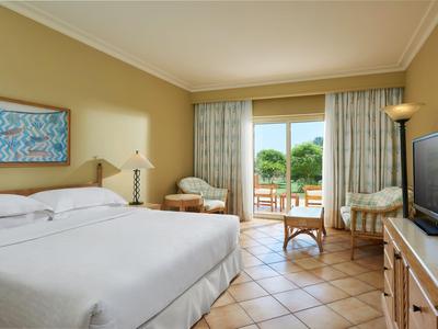 Hell eingerichtetes Hotelzimmer mit großem Bett und Blick auf Garten durch große Fenster.