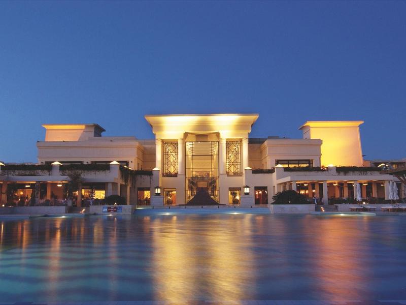 Großes, beleuchtetes Gebäude mit Säulen, davor ruhiger Wasserpool, blauer Abendhimmel.