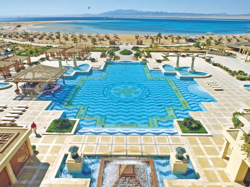 Großer blauer Resort-Pool mit Liegestühlen, Palmen und Strand im Hintergrund bei klarem Himmel.