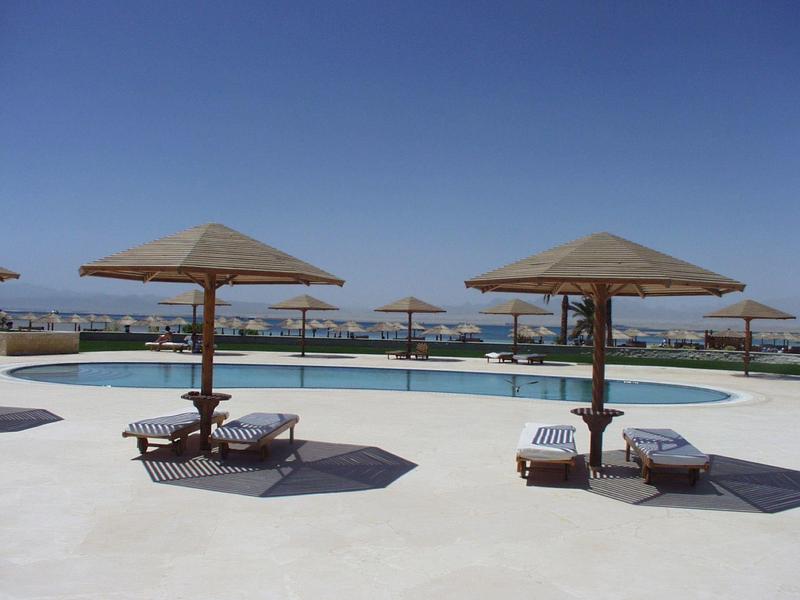 Pool mit Liegestühlen und Strohdächern vor klarem, blauem Himmel.