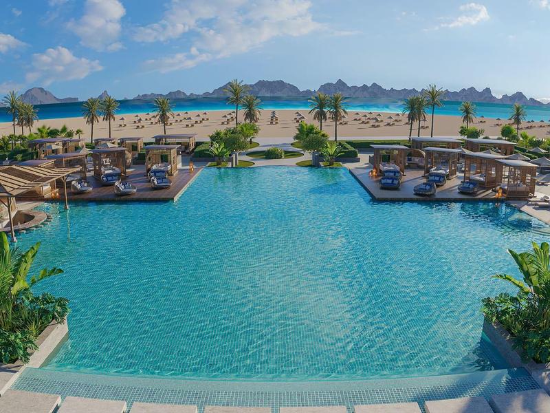 Großer blauer Pool mit Palmen und Cabanas, dahinter Strand und Berglandschaft unter blauem Himmel.