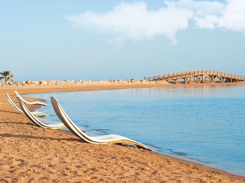Zwei weiße Sonnenliegen stehen am ruhigen Strand mit braunem Sand und blauem Meer.