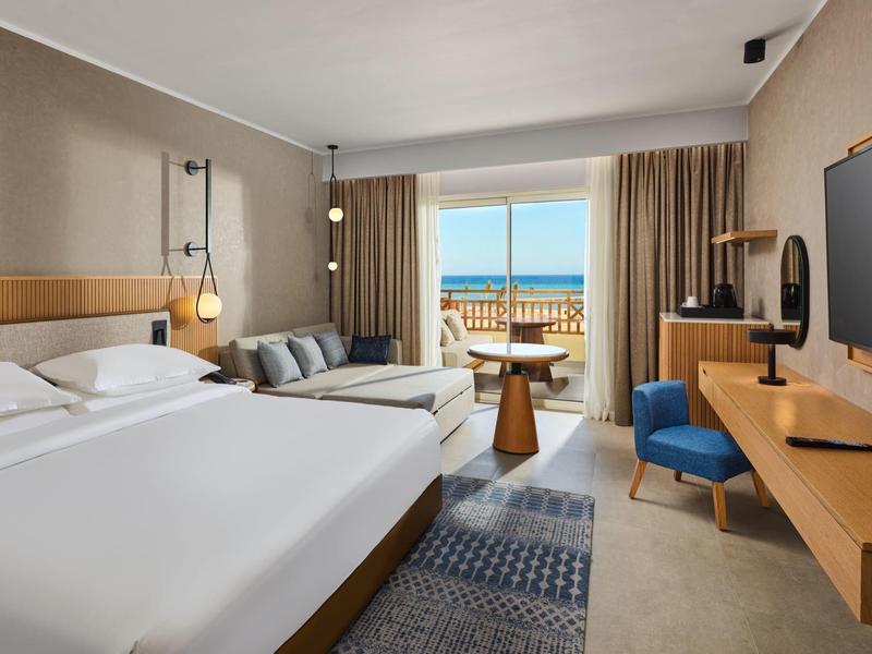 Modernes Hotelzimmer mit Doppelbett, Sofa, Schreibtisch, Meerblick und Balkon in warmen Farben.