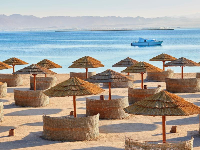 Strand mit runden Steintischen, strohgedeckten Sonnenschirmen, blauem Meer und Boot am Horizont.