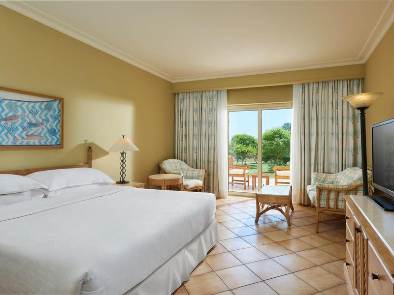 Helles Schlafzimmer mit großem Bett, Fliesenboden, TV, Sesseln und Balkon mit Blick ins Grüne.