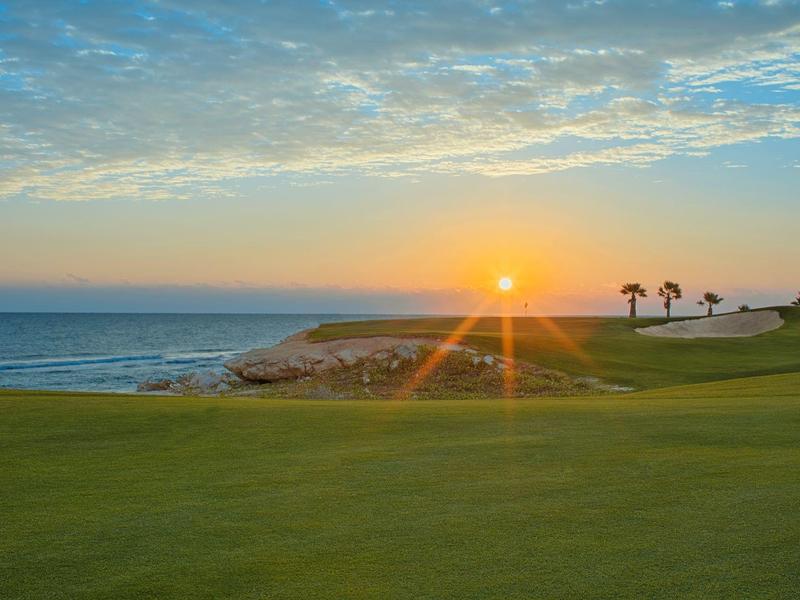 Sonnenuntergang über grünem Golfplatz mit Küste und Palmen am Horizont.