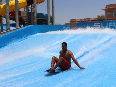 Mann surft sitzend auf blauem Wasserfluss in Wasserpark mit gelben Rutschen und klarem Himmel.