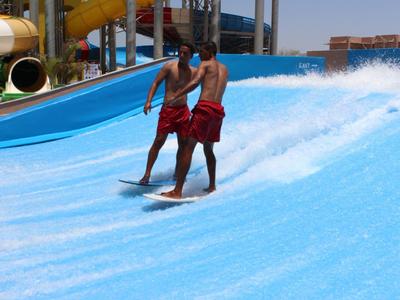 Zwei Männer surfen nebeneinander auf stehender Welle in blauer Wasseranlage mit Rutschen.