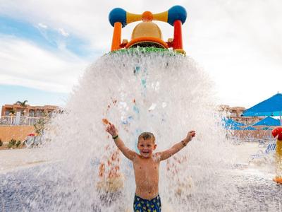 Kind steht mit ausgebreiteten Armen unter riesigem Wasserfall auf Wasserrutsche im Freizeitpark.