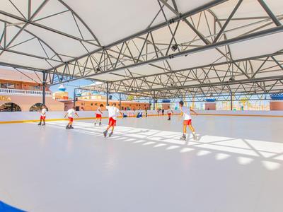 Mehrere Kinder laufen auf einer überdachten Rollhockey-Bahn mit weißen Wänden und blauem Rand.