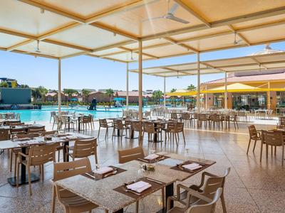 Großer Außenbereich eines Restaurants mit Holzstühlen und Tischen neben einem Pool unter Sonnensegeln.