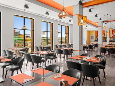 Helle Cafeteria mit großen Fenstern, schwarzen Stühlen und orangefarbenen Dekoakzenten.