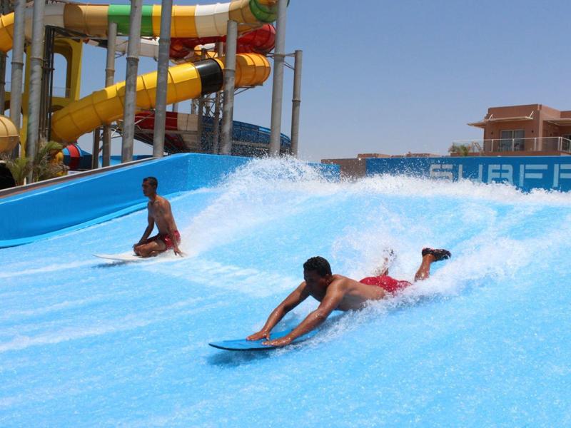 Two people surfing on artificial blue water slides at a water park on a sunny day.