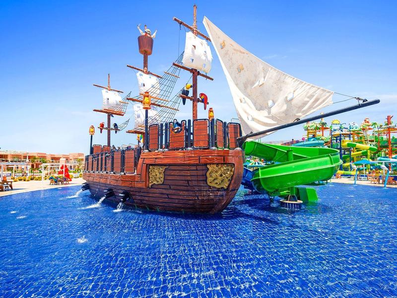 Water playground with pirate ship structure and green slides in a resort pool under blue sky.