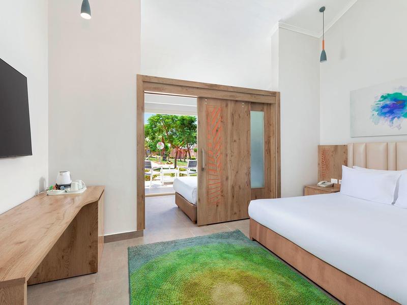 Bright hotel room with wooden furniture, a green rug, and a sliding door opening to a garden.