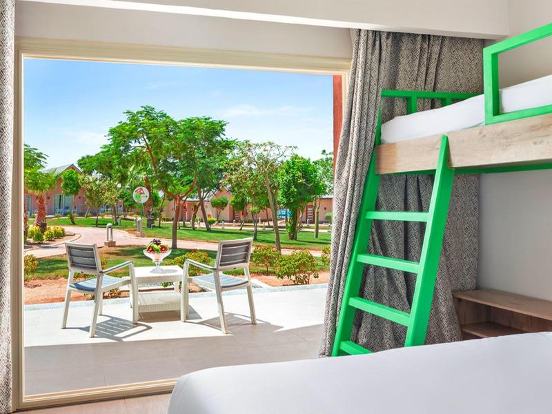 Hotel room with green bunk bed opening to patio with chairs and garden view.