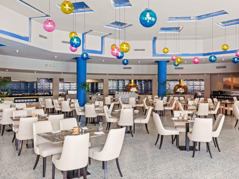 Bright, modern hotel dining area with white chairs, wooden tables, and colorful hanging decorations.