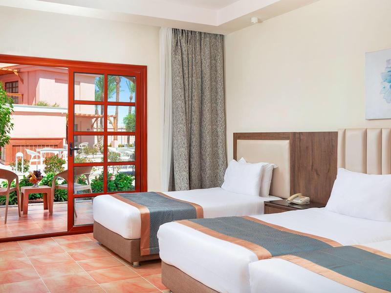 Bright hotel room with twin beds, patio doors, and outdoor seating area with greenery.