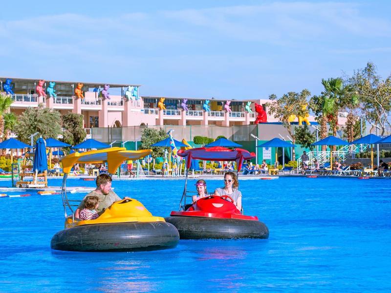 Two people ride colorful bumper boats in a blue pool in front of a hotel.