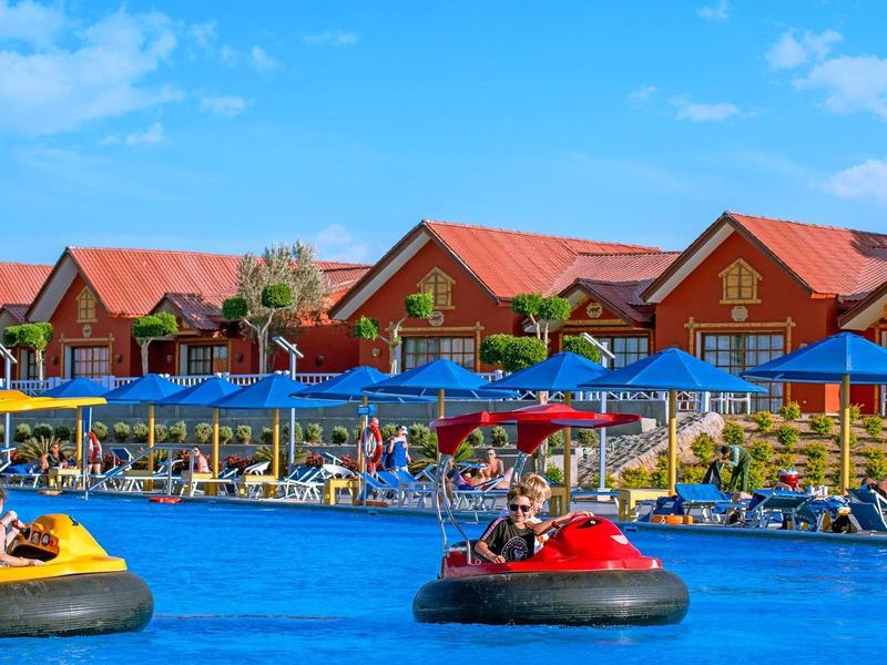 People riding bumper boats in a pool in front of wooden houses with red roofs.