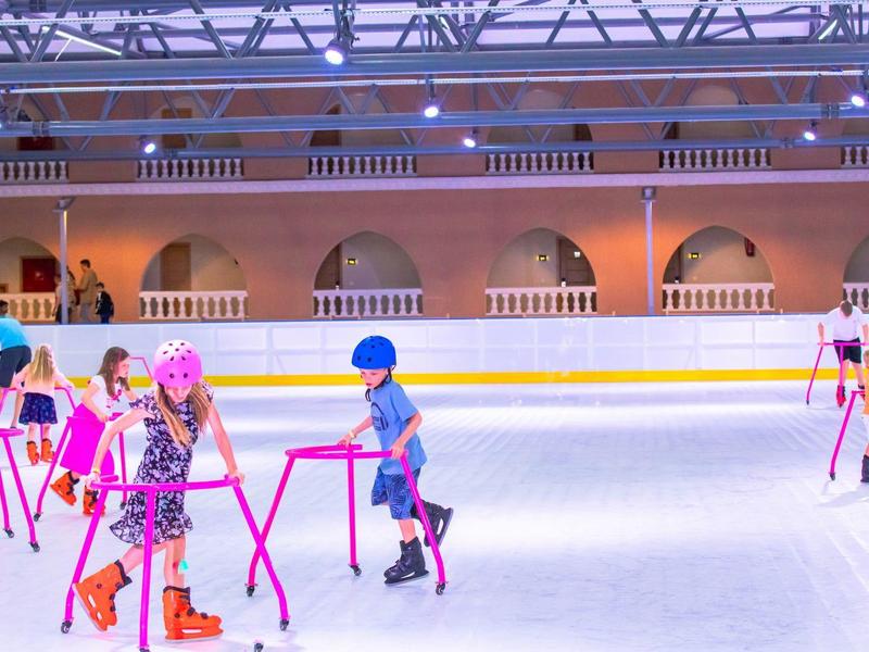 Children learning to ice skate in a bright indoor ice rink using skating aids.