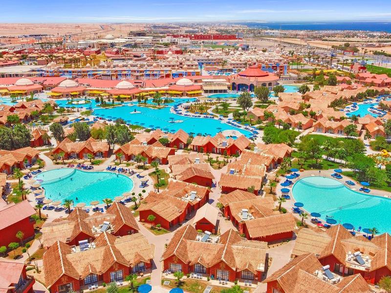 Resort with multiple pools and red-roofed villas in a desert landscape near the coast.