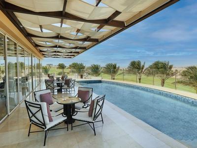 Terrazza coperta con area salotto accanto a una piscina con vista su un prato e alberi.
