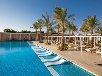 Piscina esterna moderna con lettini bassi e palme in un resort alberghiero sotto un cielo limpido.