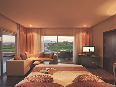 Hotel room with large bed, sofa, desk, TV, and windows overlooking a golf course at sunset.