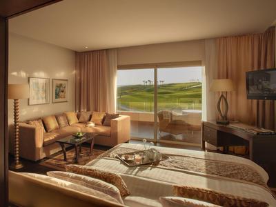 Cozy hotel room with beige décor, a sofa, bed, TV, and a balcony overlooking a green golf course.