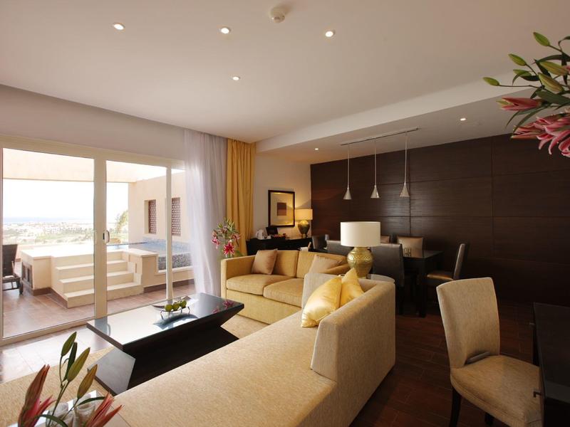 Modern hotel suite with beige sofas, dark wood accents, and a balcony overlooking water.