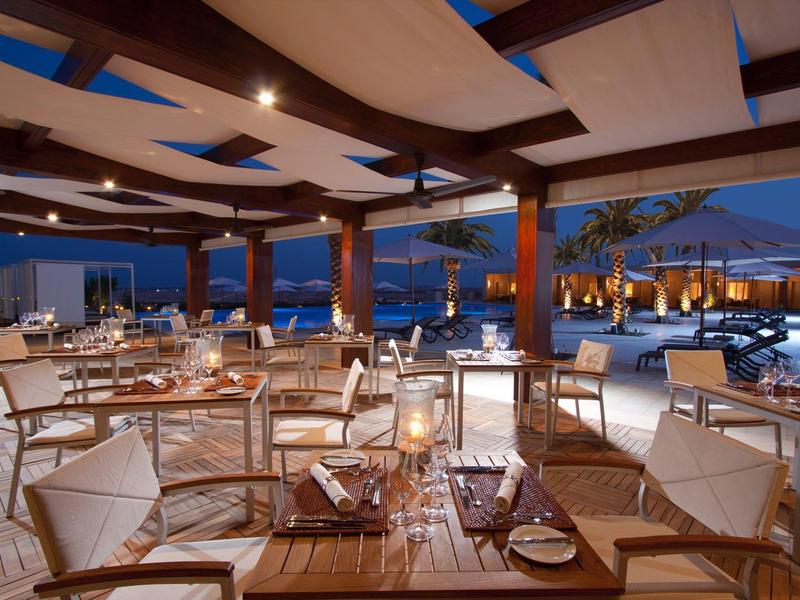 Outdoor hotel restaurant with wooden furniture and pool at dusk under a canopy.