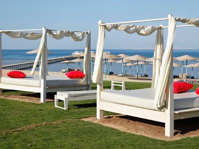 White daybeds with red cushions on grass by the sea and beach umbrellas.