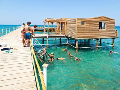 Mehrere Personen schwimmen und entspannen sich an einem Holzsteg mit einem über dem Wasser gebauten Haus.